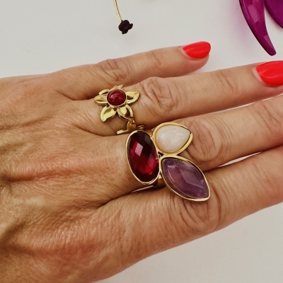 Mão com anéis dourados e pedras coloridas, unhas vermelhas, fundo branco com flores roxas