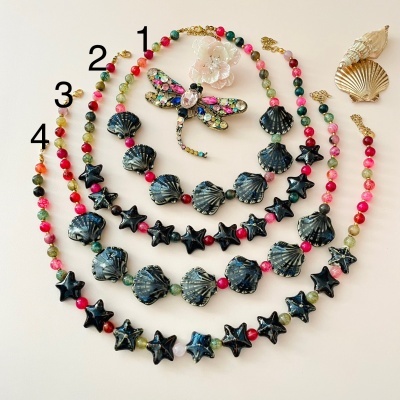 Quatro colares com contas coloridas e pérolas negras, broche de libélula multicolorido e duas conchas no fundo.