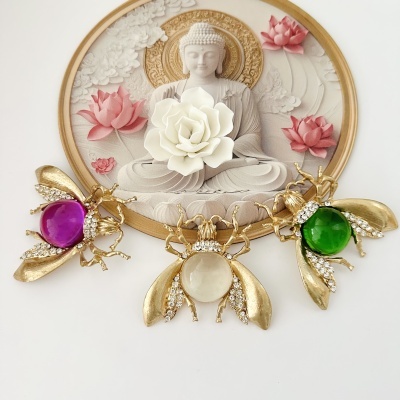 Broches de abelha dourada com pedras roxa, branca e verde, frente a placa decorativa com Buda e flores de lótus.