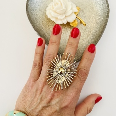 Mão com anel decorativo dourado e unhas vermelhas sobre prato prateado em forma de coração com flor branca