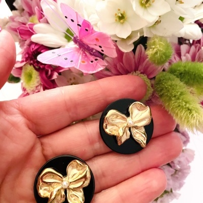 Brincos redondos pretos com laços dourados e pérola em frente a flores e borboleta rosa