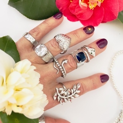 Mão com vários anéis prateados e flores coloridas ao fundo
