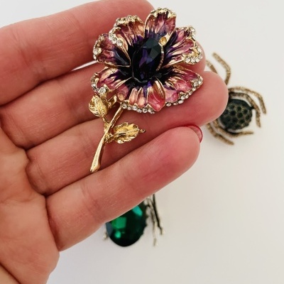 Broche de flor roxa com pedra azul escura e detalhes dourados, segurado por mão com unhas vermelhas