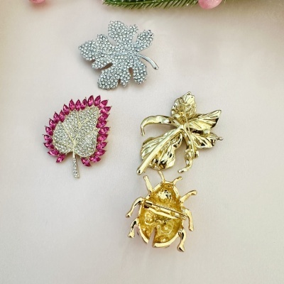 Três broches decorativos forma folhas e inseto, prateados com cristais e dourado, sobre superfície clara.