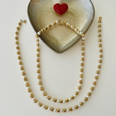 Colar e pulseira dourados com contas brancas e douradas ao lado de uma travessa metálica cinza em forma de coração com um pequeno coração vermelho no centro