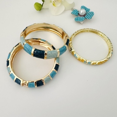 Pulseiras douradas com padrões azuis e flor branca ao fundo