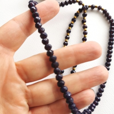 Colar preto com contas facetadas e detalhes dourados, segurado por mão, fundo branco e flores cor-de-rosa