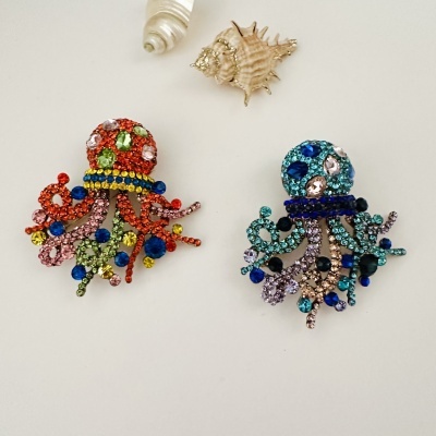 Dois broches coloridos em forma de polvo com pedras brilhantes e duas conchas ao fundo em superfície branca
