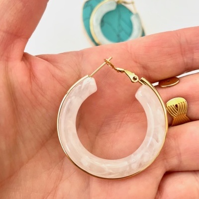 Brinco de argola dourada com resina branca sendo segurado numa mão