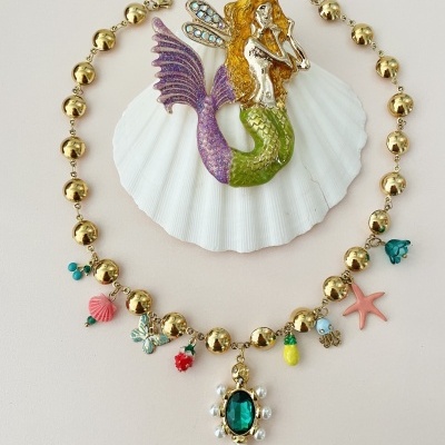 Colar dourado com pendentes coloridos em fundo claro com concha e figura de sereia.