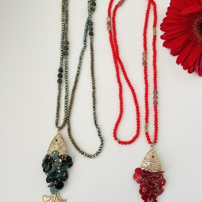 Duas colares longos em contas pretas e vermelhas com pendentes de peixe dourados, sobre fundo branco com flor vermelha ao lado.