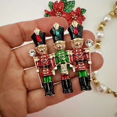 Broche com três soldados coloridos e brilhantes segurando bastões, em metal esmaltado e pedras decorativas.