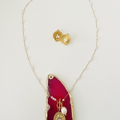 Colar dourado com pingente oval decorado com pérolas e brincos dourados em forma de concha, disposto sobre placa de ágata vermelha.