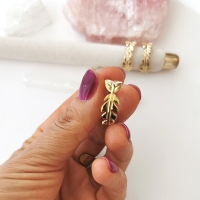 Mão segurando anel dourado com design de folhas com unhas pintadas de roxo e pedras decorativas ao fundo