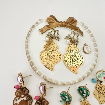 Brincos dourados com padrão vazado e pérolas num prato branco com laço dourado
