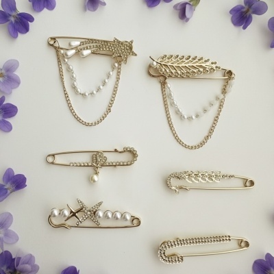 Alfinetes dourados decorativos com pérolas e cristais sobre fundo branco com flores roxas