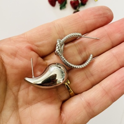 Dois brincos em prata, um em meia-lua texturizado e outro em forma de pimento, segurados na mão com fundo branco e morangos ao fundo.