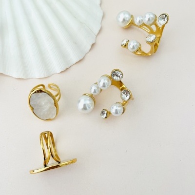 Quatro anéis dourados com pérolas, pedras e design minimalista junto a uma concha branca