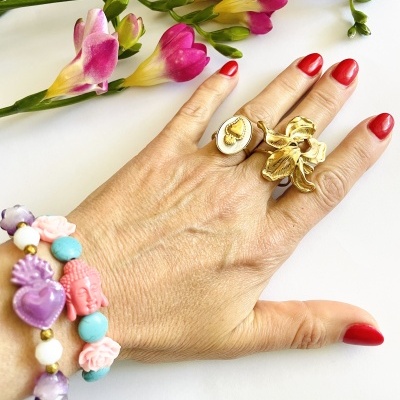 Mão feminina com pulseiras coloridas e anéis dourados junto a flores coloridas sobre fundo branco.