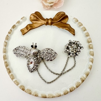 Broche prateado com strass em forma de abelha e círculo floral em base branca com laço dourado
