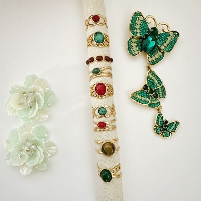 Anéis dourados com pedras coloridas num suporte branco, brincos florais verdes claros com pérolas e brincos borboleta verdes e dourados.