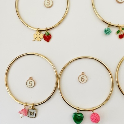 Pulseiras douradas com pendentes coloridos e marcadores numerados de 3 a 6 sobre fundo branco