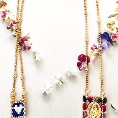 Dois colares dourados com pendentes quadrados e pedras, sobre fundo claro com flores