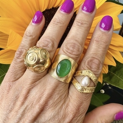 Mão com três anéis dourados e unhas pintadas de rosa forte frente a um girassol amarelo