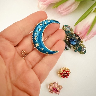Broche azul e dourado em forma de lua com estrelas, broches coloridos e flores rosas ao fundo