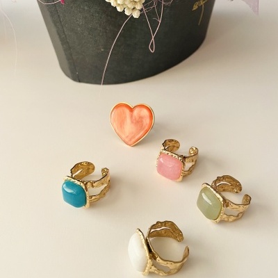 Anéis dourados com pedras coloridas e anel coração laranja sobre superfície branca com flores ao fundo