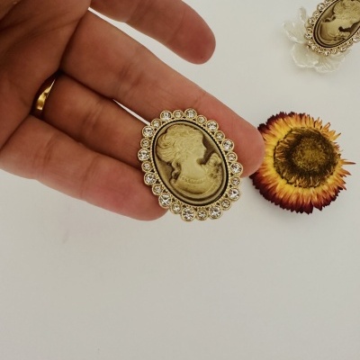 Broches vintage ovais dourados com perfil feminino e brilhantes, mão segurando broche e flor seca ao lado