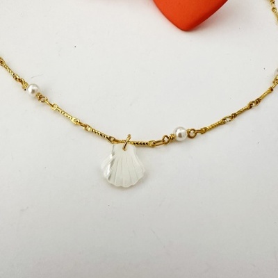 Colar dourado com pérolas brancas e pendente de concha, corações vermelhos ao fundo.