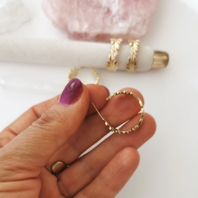 Mão segurando brinco dourado com anéis dourados e pedra rosa ao fundo