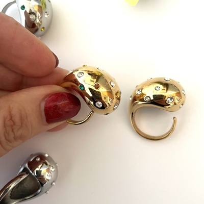 Anéis metálicos dourado e prateado com pedras coloridas e brilhantes em fundo branco com mão e flor amarela