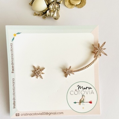 Brincos dourados em forma de estrelas com brilhantes pequenos sobre cartão branco com logotipo e contatos.