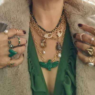 Mãos femininas com anéis e vários colares dourados, com casaco de pelo bege e blusa verde