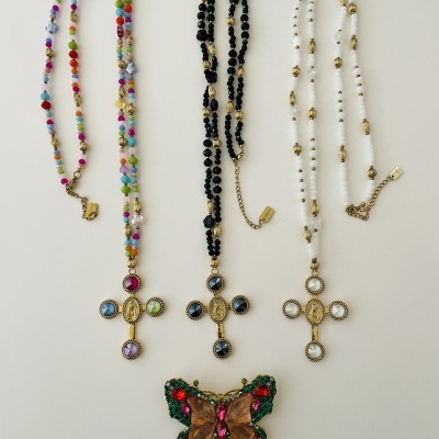 Três colares com cruzes douradas e pedras coloridas e um broche de borboleta colorida sobre fundo branco