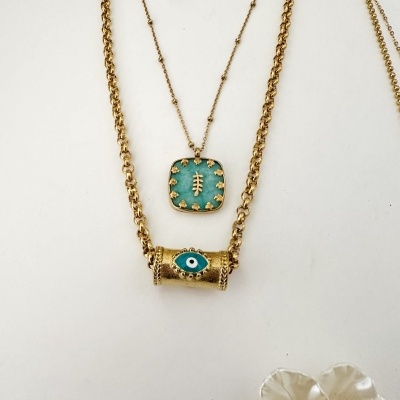Duas colares dourados com pendentes azuis e detalhes dourados, sobre fundo branco com flor decorativa.