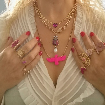 Mãos femininas com anéis dourados e rosa e colares dourados com pendentes coloridos sobre blusa branca plissada