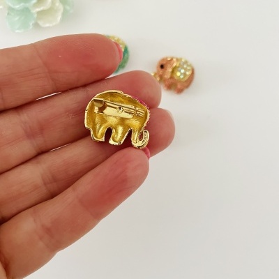 Alfinete elefante dourado nas mãos com alfinetes coloridos e flores ao fundo