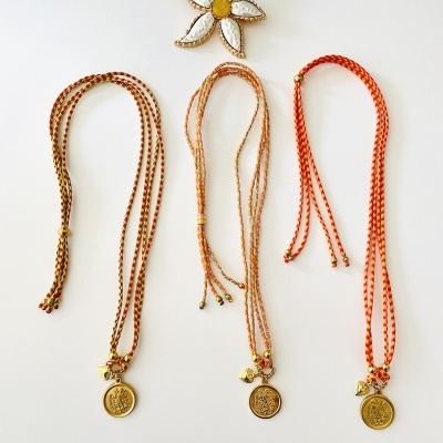 Três colares trançados com pendentes dourados sobre fundo branco e uma flor decorativa