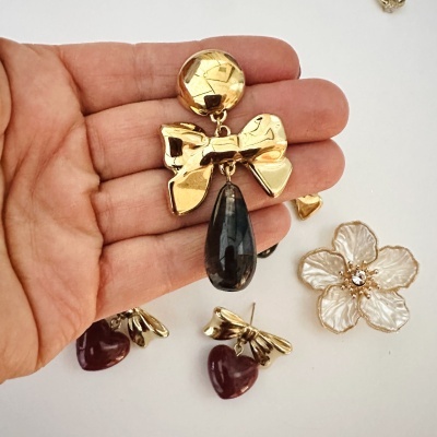 Brincos de metal dourado com laço e pendente cinza, e coração vermelho com laço metal dourado, junto com broche de flor branca