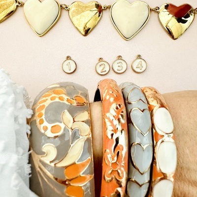 Pulseira de corações dourados e brancos com medalhas numeradas e pulseiras esmaltadas em tons de cinza, laranja, dourado e branco no pulso