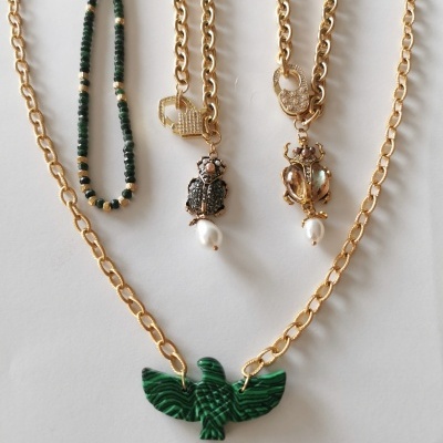 Quatro colares com correntes douradas e pedentes, incluindo um pendente verde em forma de pássaro e colar de contas verdes e douradas.