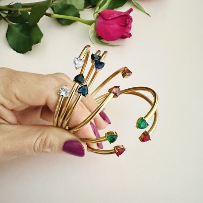 Pulseiras douradas com pedras em forma de coração em várias cores seguras por mão feminina com unhas roxas com uma rosa vermelha ao fundo