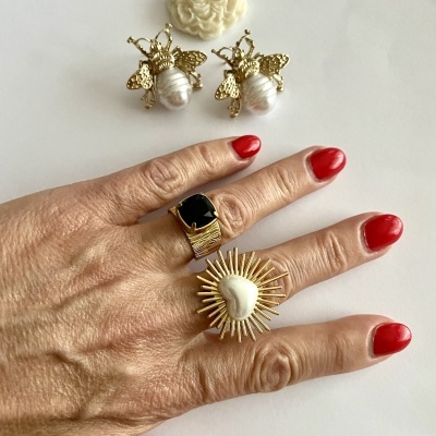 Mão com anéis dourados e unhas vermelhas junto a brincos dourados e broche creme