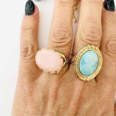 Mão com dois anéis grandes com pedras ovais rosa e azul e unhas pretas com brilhos.