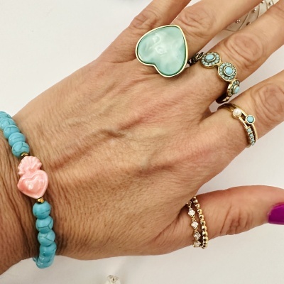 Mão com anéis dourados com pedras turquesa, pulseira azul e rosa, unhas roxas sobre fundo branco