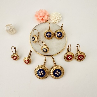 Brincos pendentes redondos dourados com detalhes azuis e vermelhos e flores decorativas numa superfície branca.