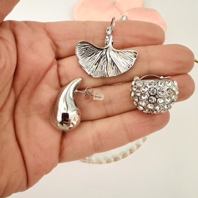 Três brincos prateados em formato de folha, gota e com pedras brilhantes na mão